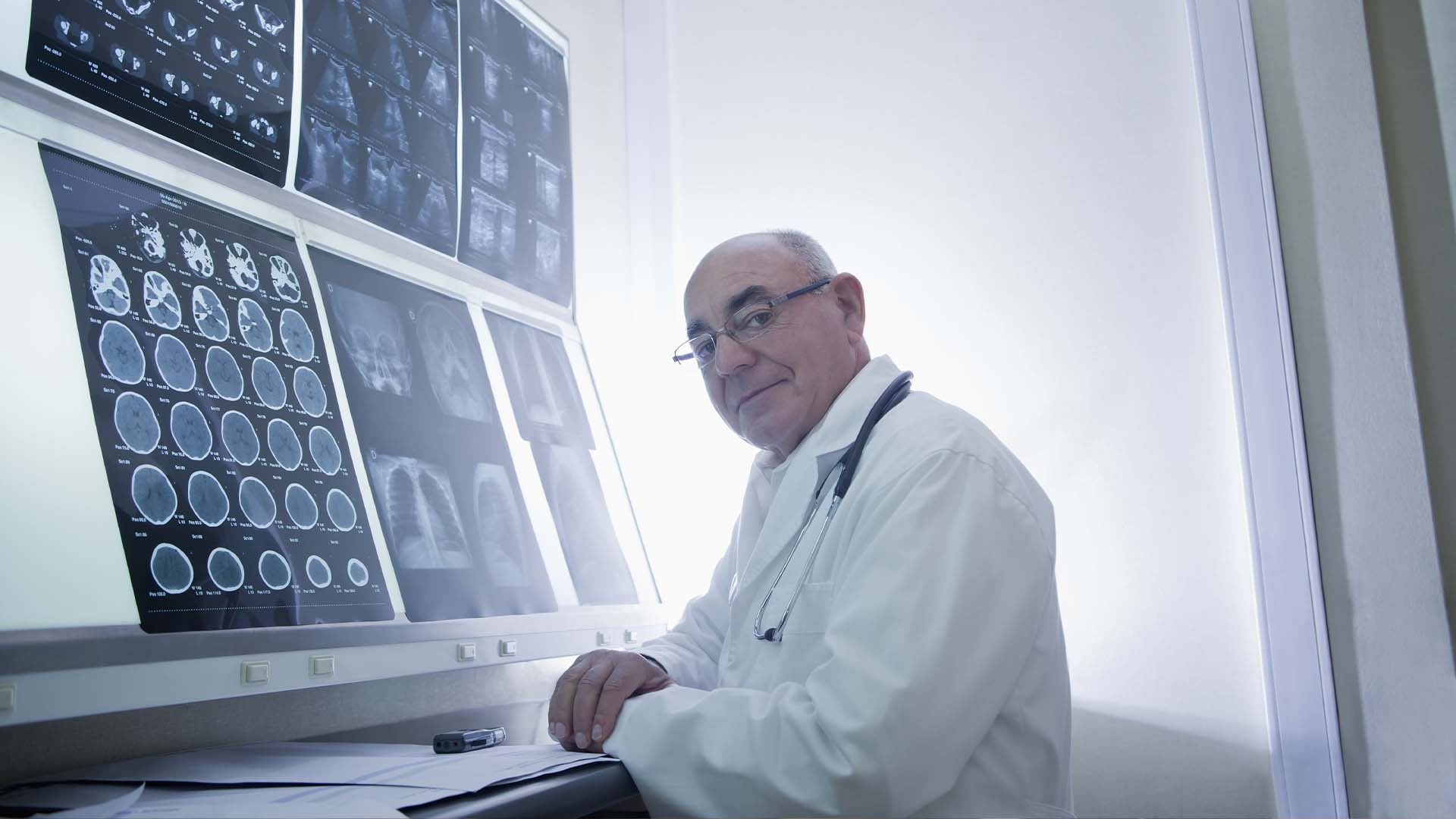 Male neurologist in white coat with glasses and stethoscope reviewing multiple brain CT and MRI scan images displayed on illuminated medical monitors in radiology room