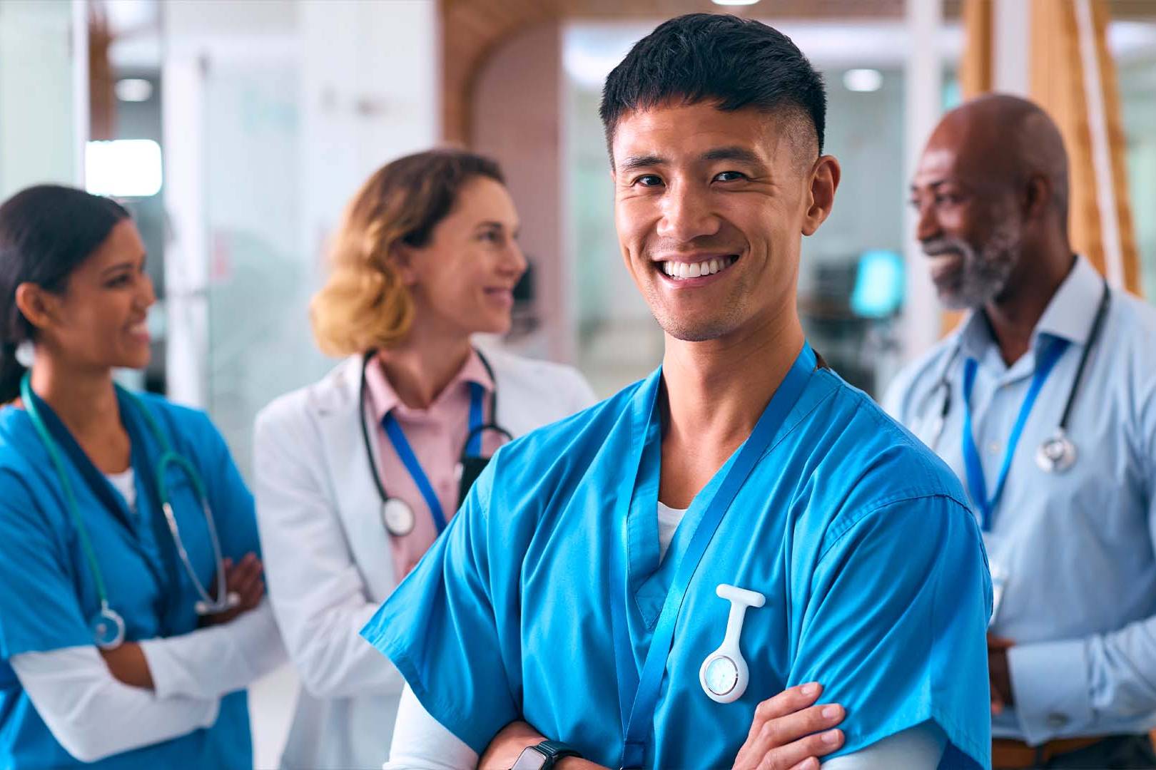 Smiling Asian male nurse practitioner in blue scrubs with stethoscope standing confidently with diverse healthcare team in bright modern hospital corridor
