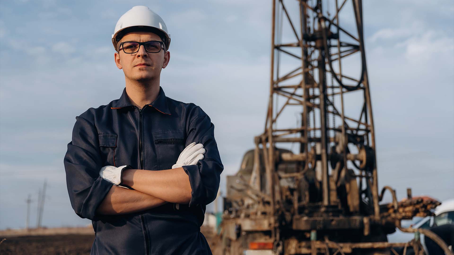 Confident oil rig engineer in hard hat and workwear standing arms crossed at drilling site