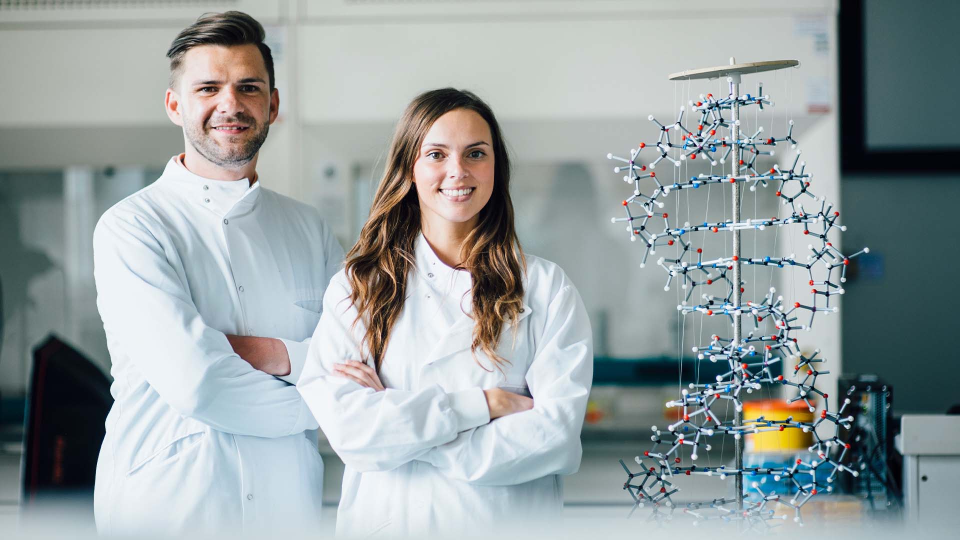 Male and female scientists in white lab coats standing confidently with arms crossed beside a large molecular structure model in a bright modern laboratory