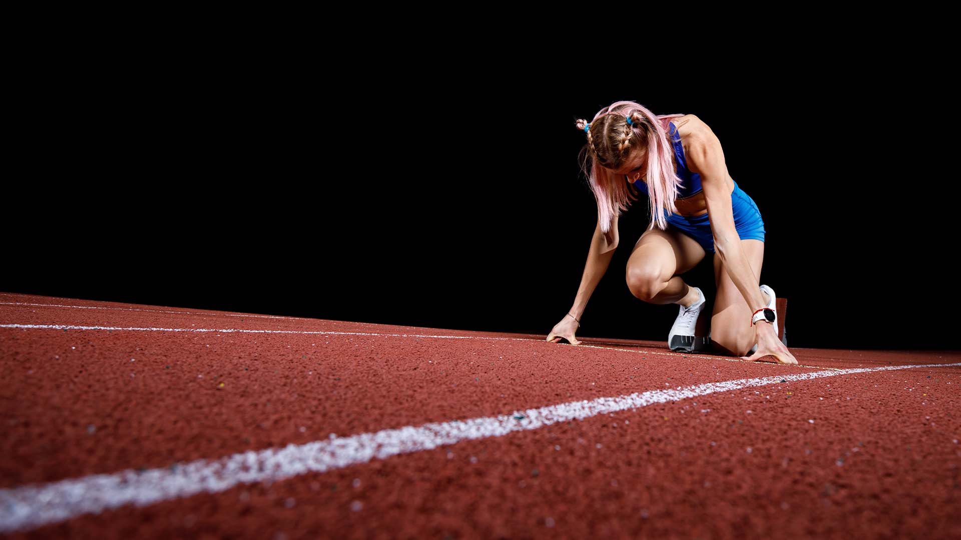 Female athlete in blue athletic wear crouched down on red running track with head bowed in moment of exhaustion or contemplation against black background