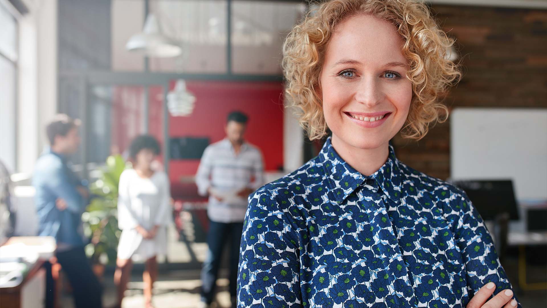 Smiling woman with curly blonde hair in blue floral patterned blouse standing confidently with arms crossed in modern office with colleagues meeting in background