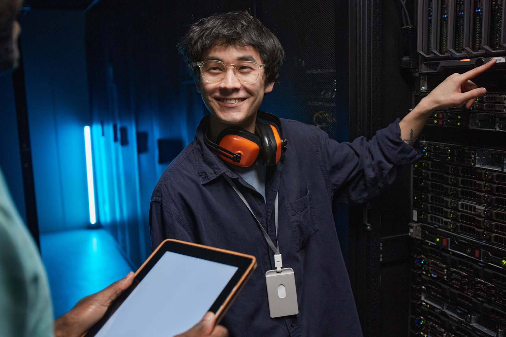 Tech worker with headphones and ID badge gesturing at server racks in blue-lit data center while colleague holds tablet
