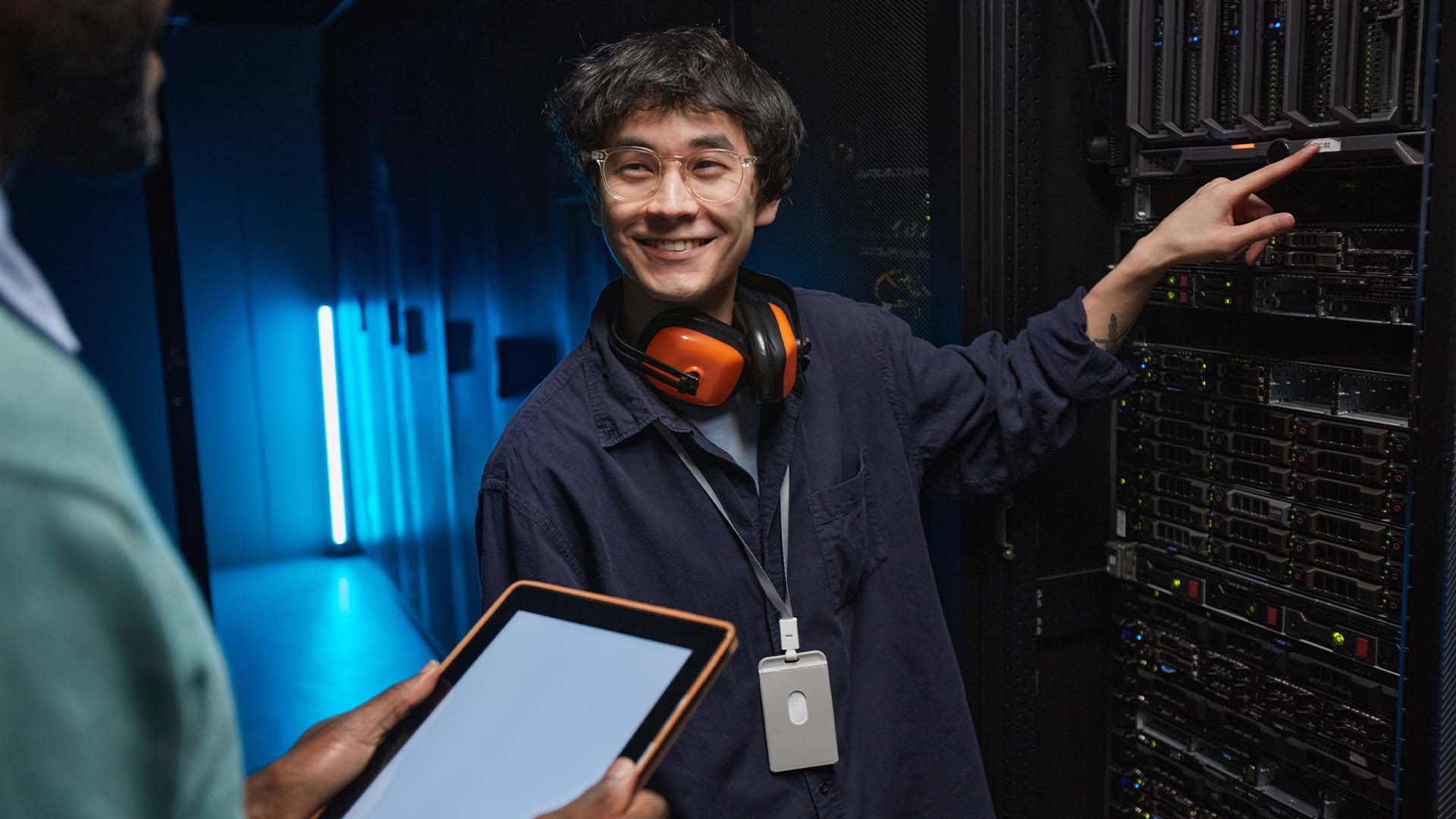 Tech worker with headphones and ID badge gesturing at server racks in blue-lit data center while colleague holds tablet