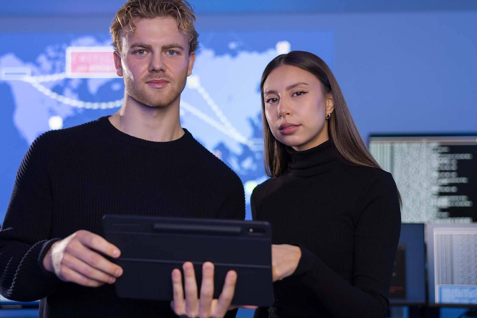 Two tech workers in black clothing holding tablet in front of global data screens with serious expressions