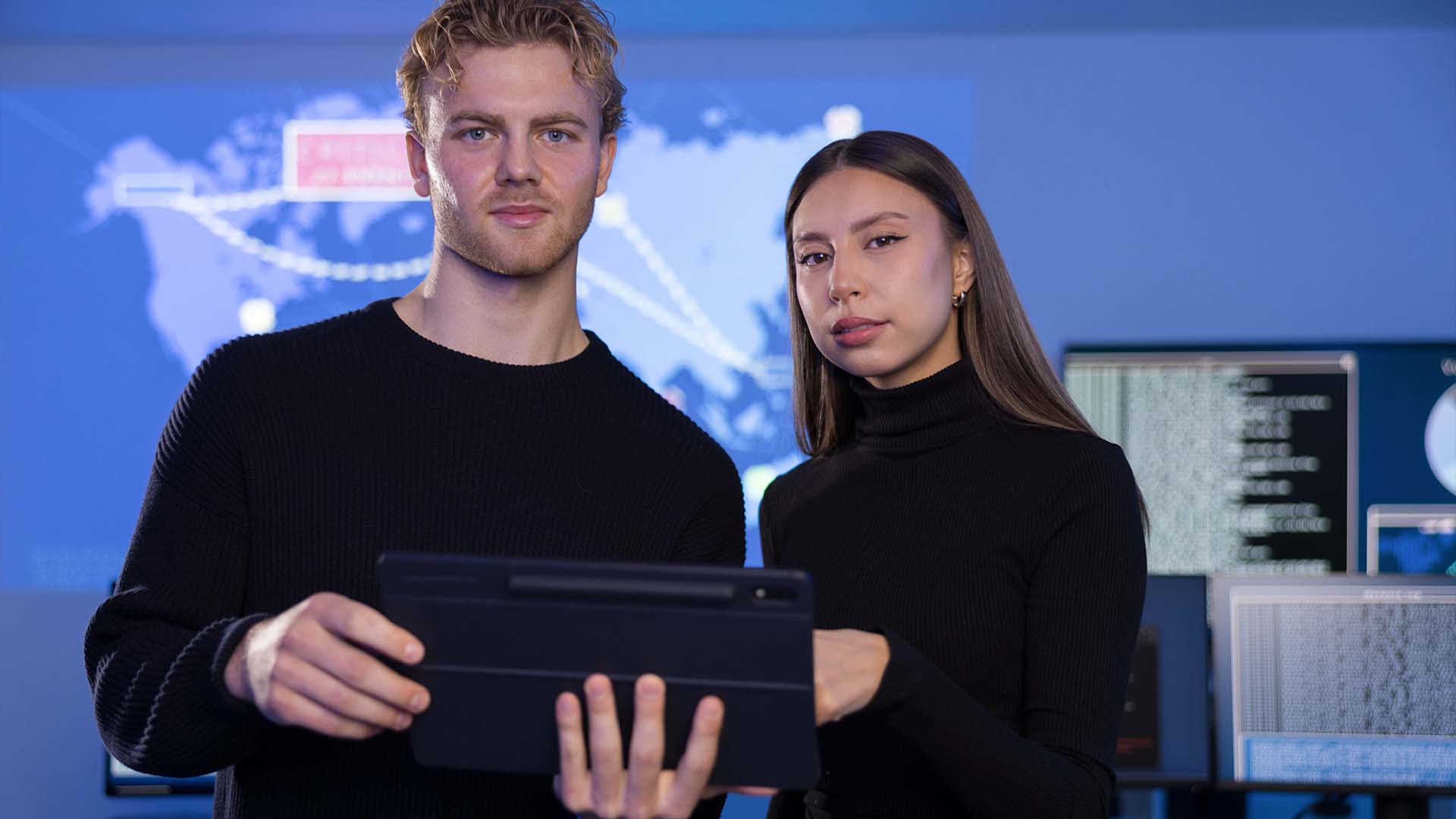 Two tech workers in black clothing holding tablet in front of global data screens with serious expressions