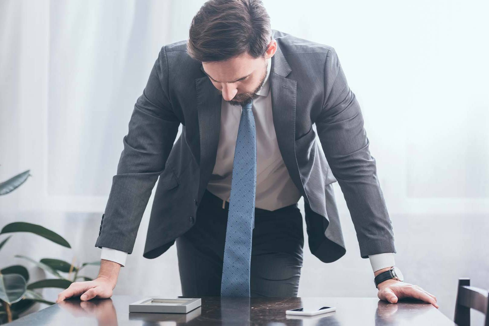 Exhausted executive in suit leaning on desk in frustration in bright office