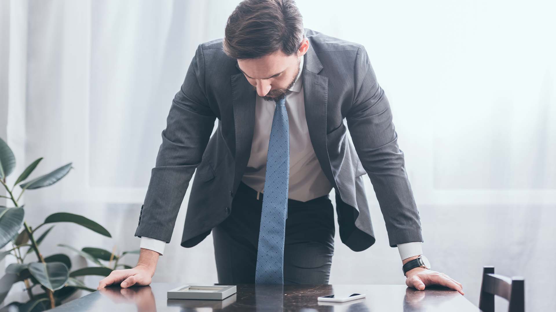 Exhausted executive in suit leaning on desk in frustration in bright office