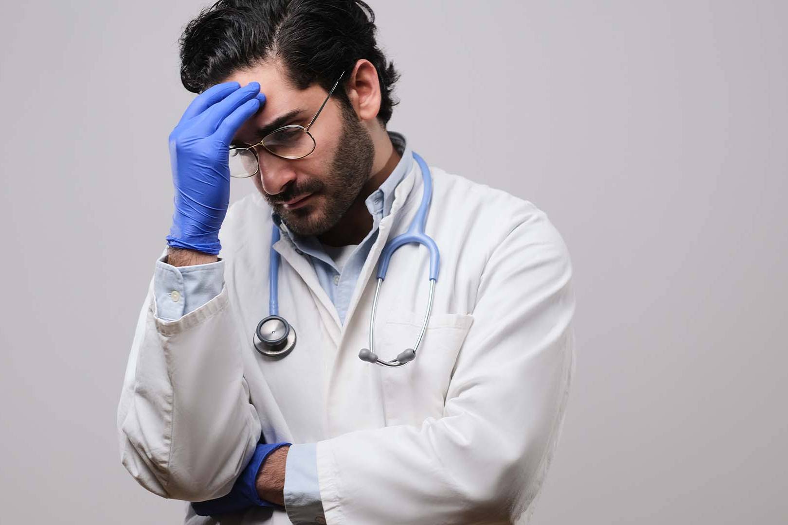 Stressed male doctor in lab coat and gloves holding forehead in frustration