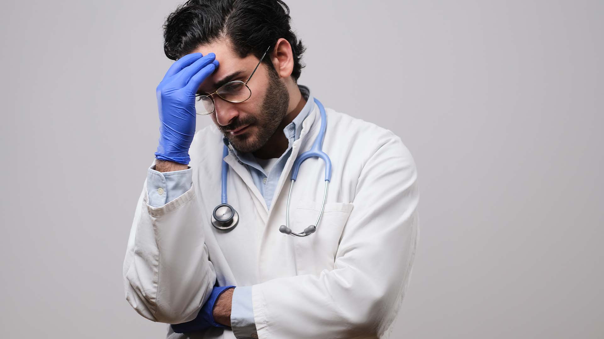 Stressed male doctor in lab coat and gloves holding forehead in frustration
