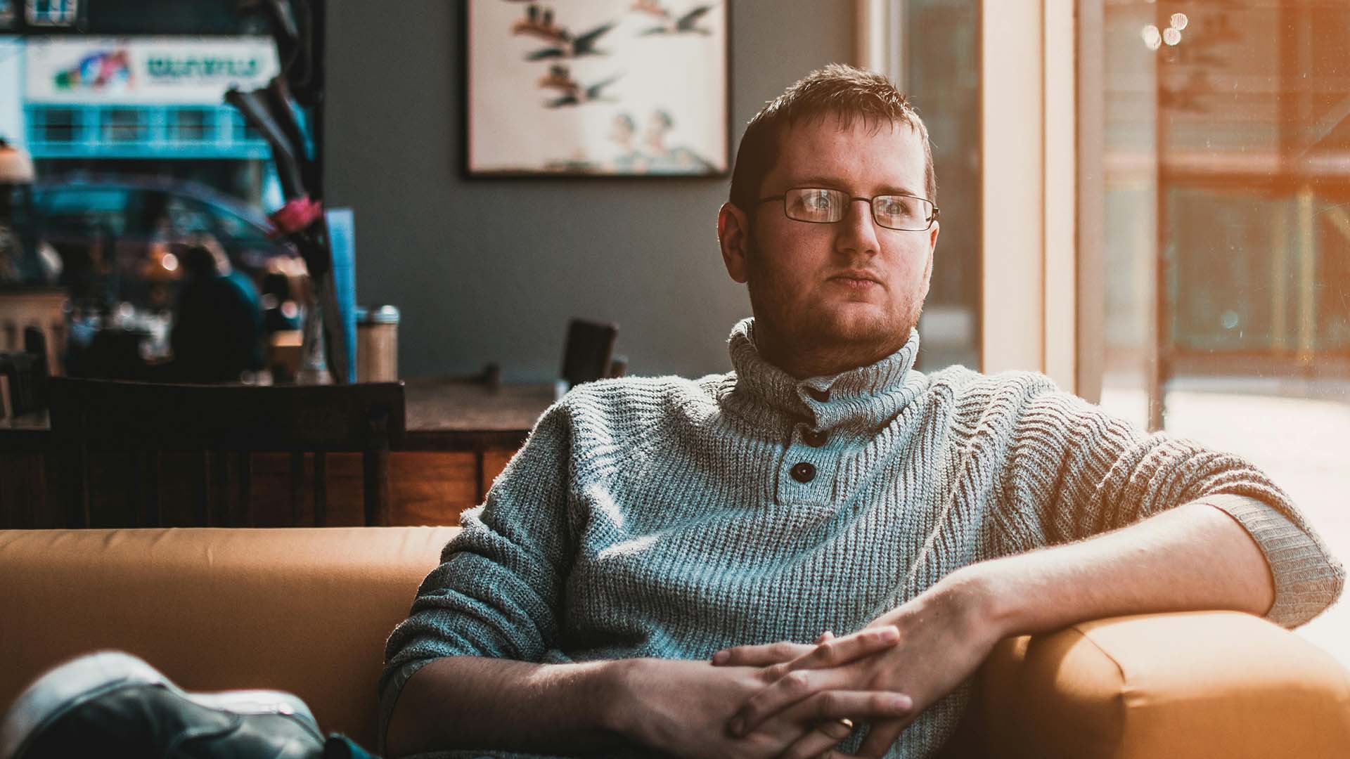 A young man in a knit turtleneck sweater sitting on a leather couch with his hands folded, gazing off thoughtfully