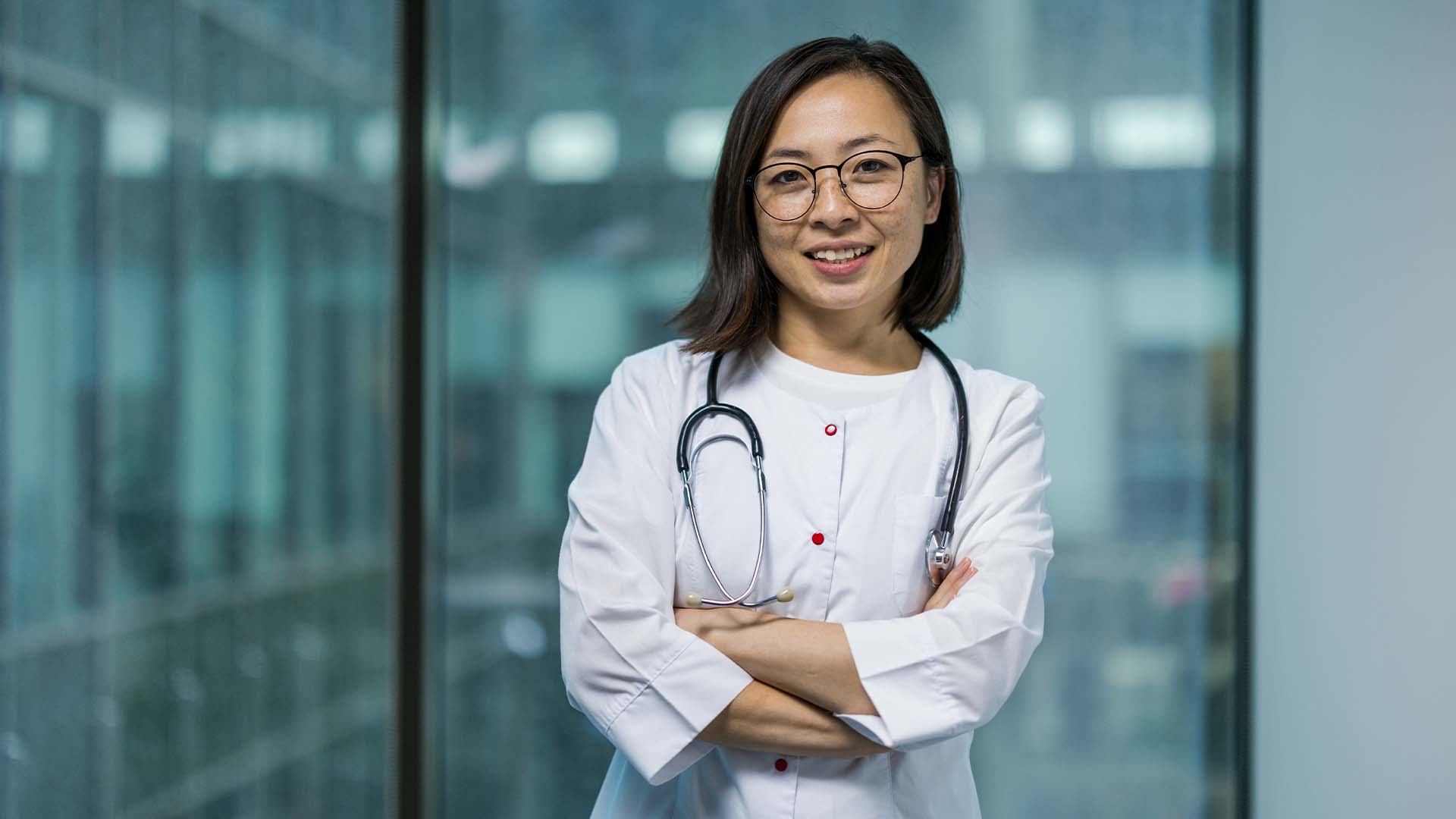 Confident female physician in a white coat with a stethoscope standing in a modern office.