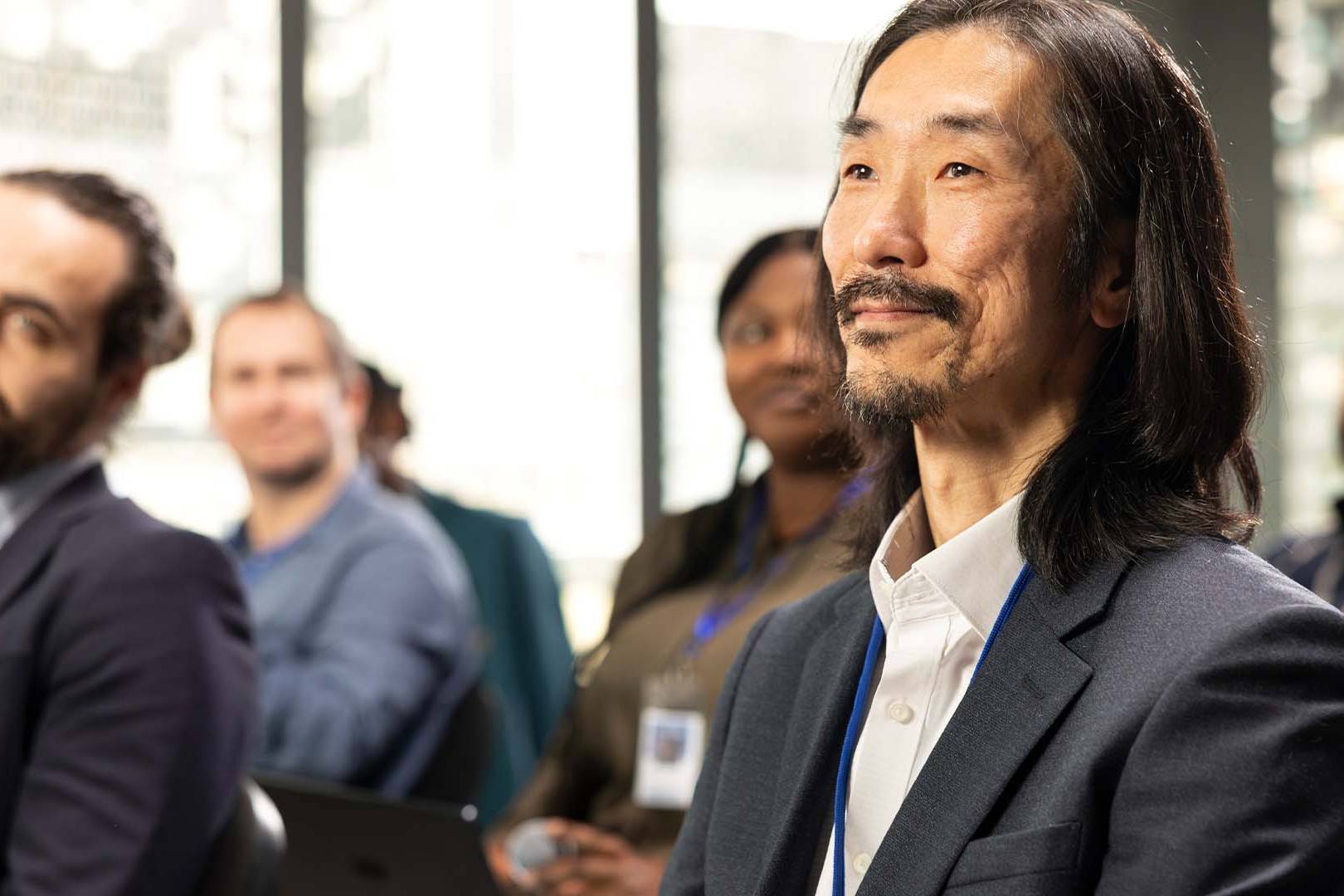 Asian man with long dark hair in a navy blazer and conference badge listening attentively at a corporate board meeting or conference
