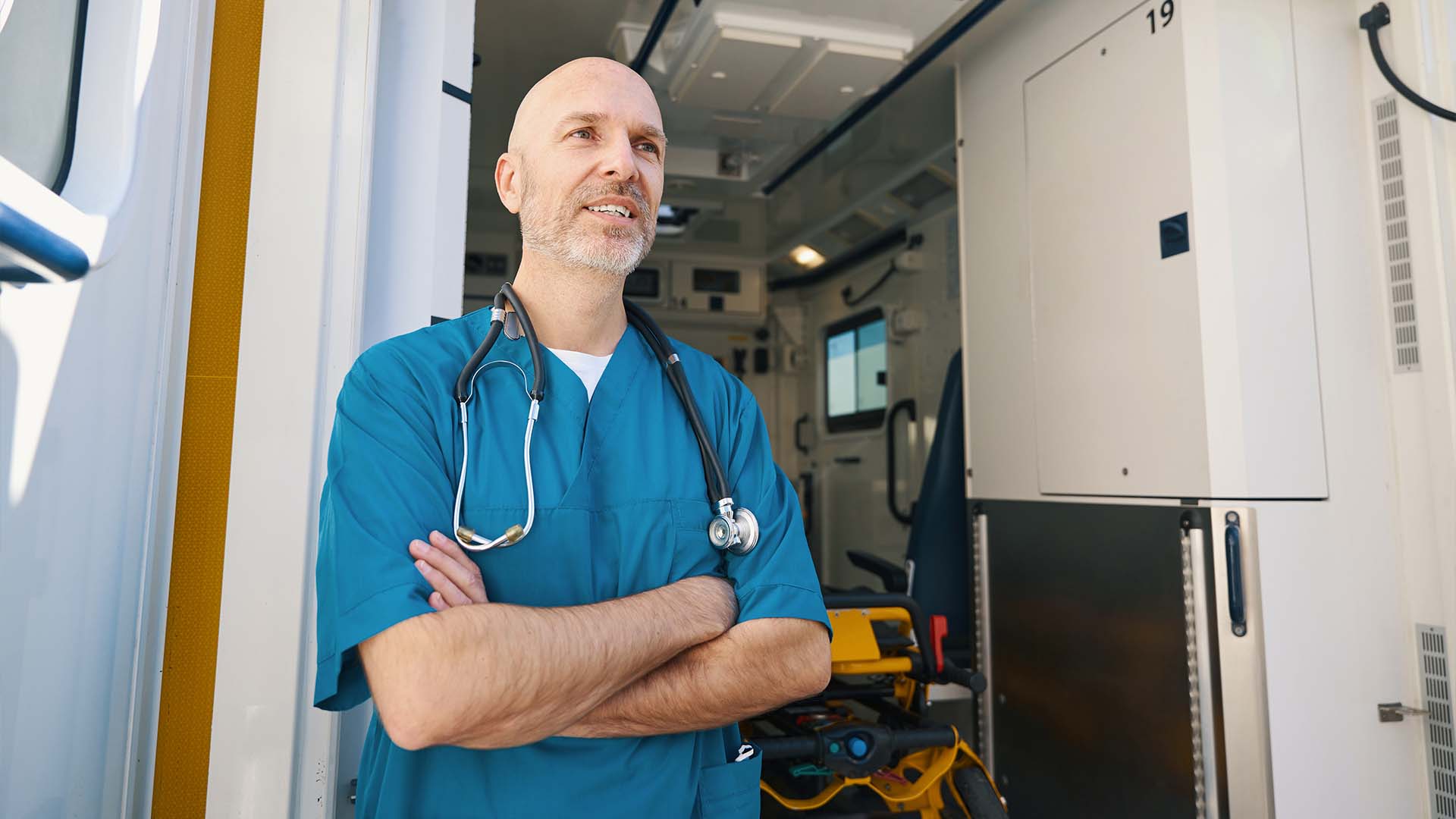 Confident male emergency physician in blue scrubs with stethoscope standing with arms crossed at ambulance entrance