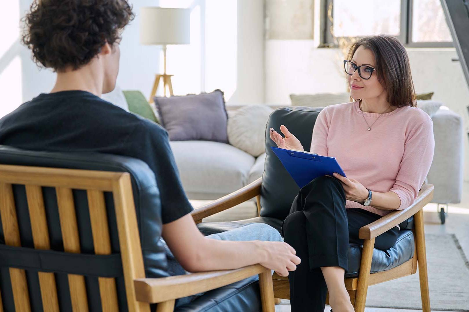 Therapist with glasses holding a clipboard while speaking with a client during a one-on-one counseling session