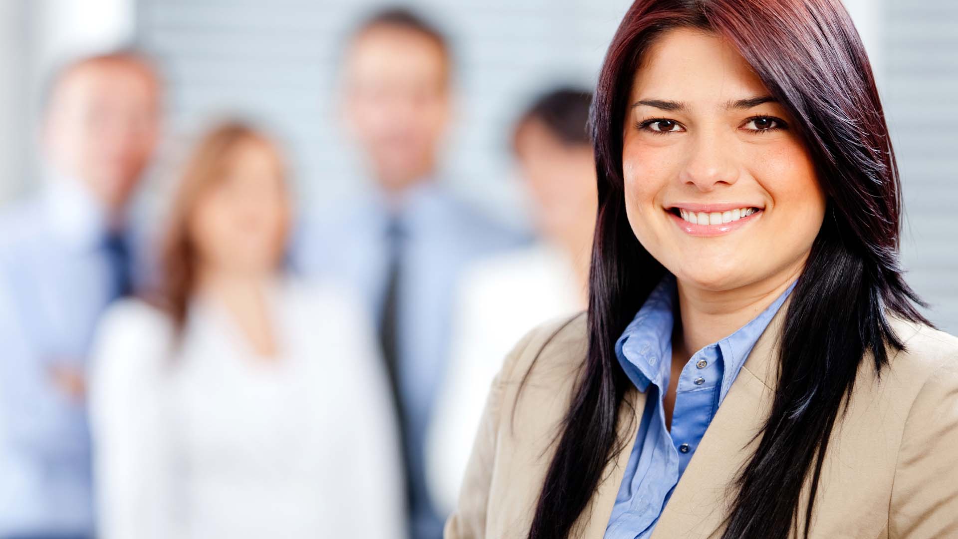 Professional woman with dark hair smiling confidently in a business setting with colleagues in the background