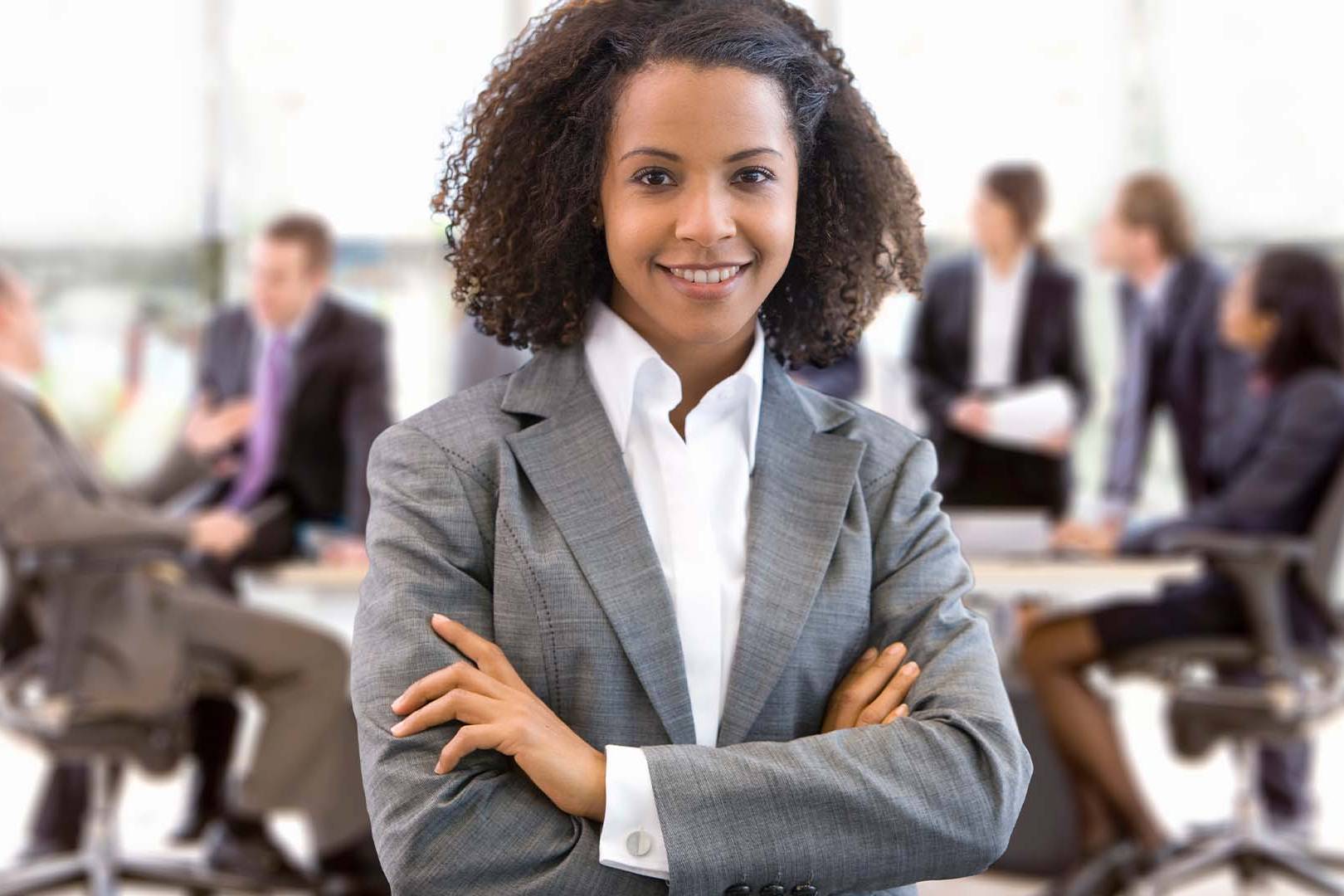 Confident businesswoman with arms crossed standing in a corporate office with colleagues meeting in the background