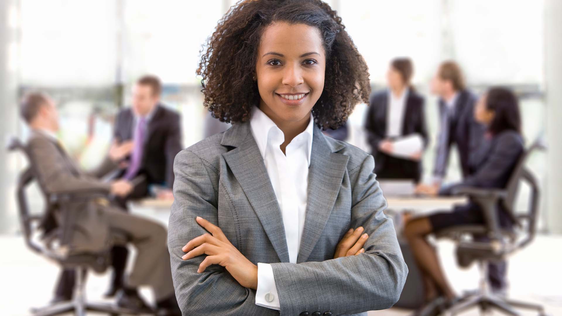 Confident businesswoman with arms crossed standing in a corporate office with colleagues meeting in the background