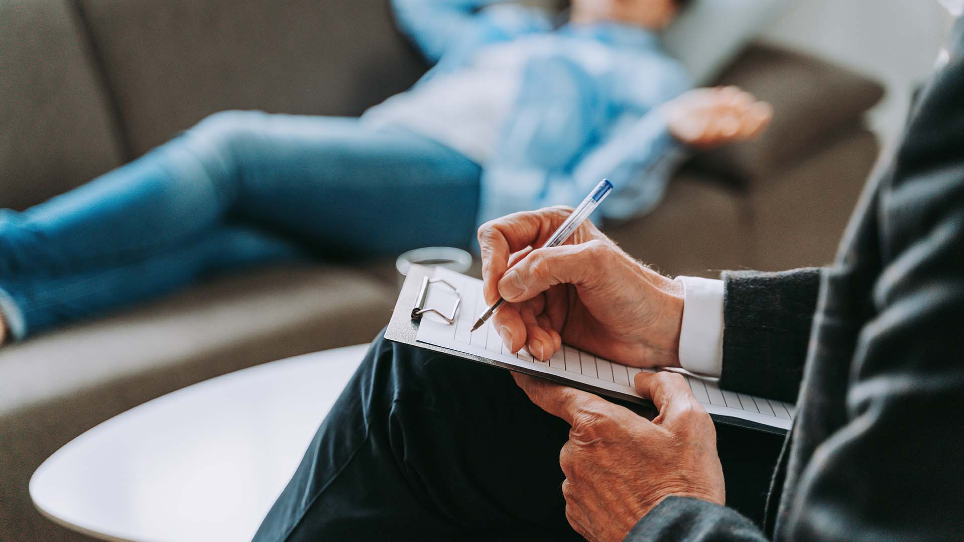 Close-up of a therapist taking notes on a clipboard during a session with a client reclining on a couch