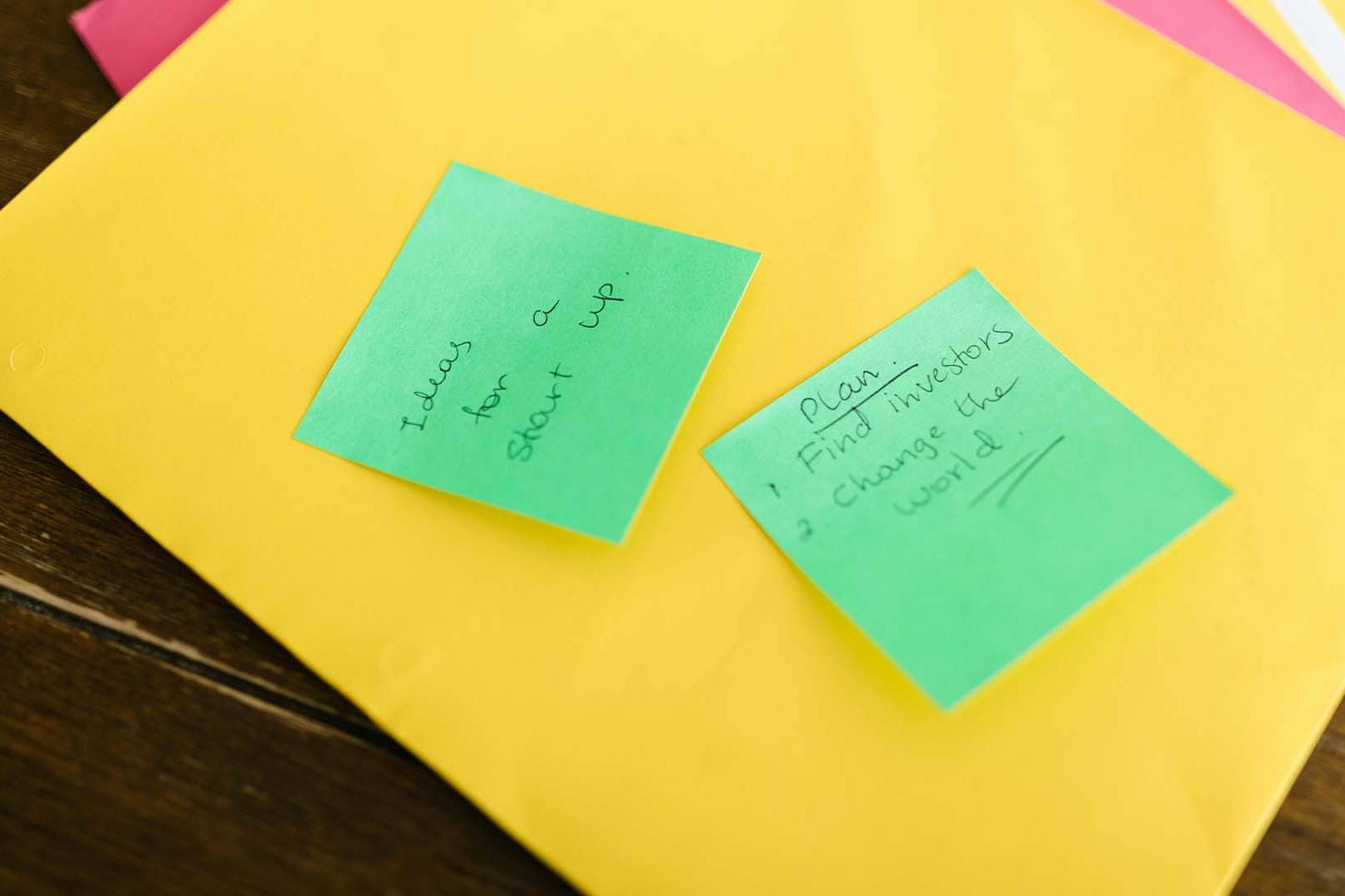 Two green sticky notes on a yellow folder with handwritten startup plans and ideas on a dark wooden desk