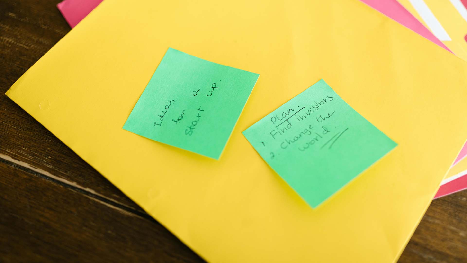 Two green sticky notes on a yellow folder with handwritten startup plans and ideas on a dark wooden desk