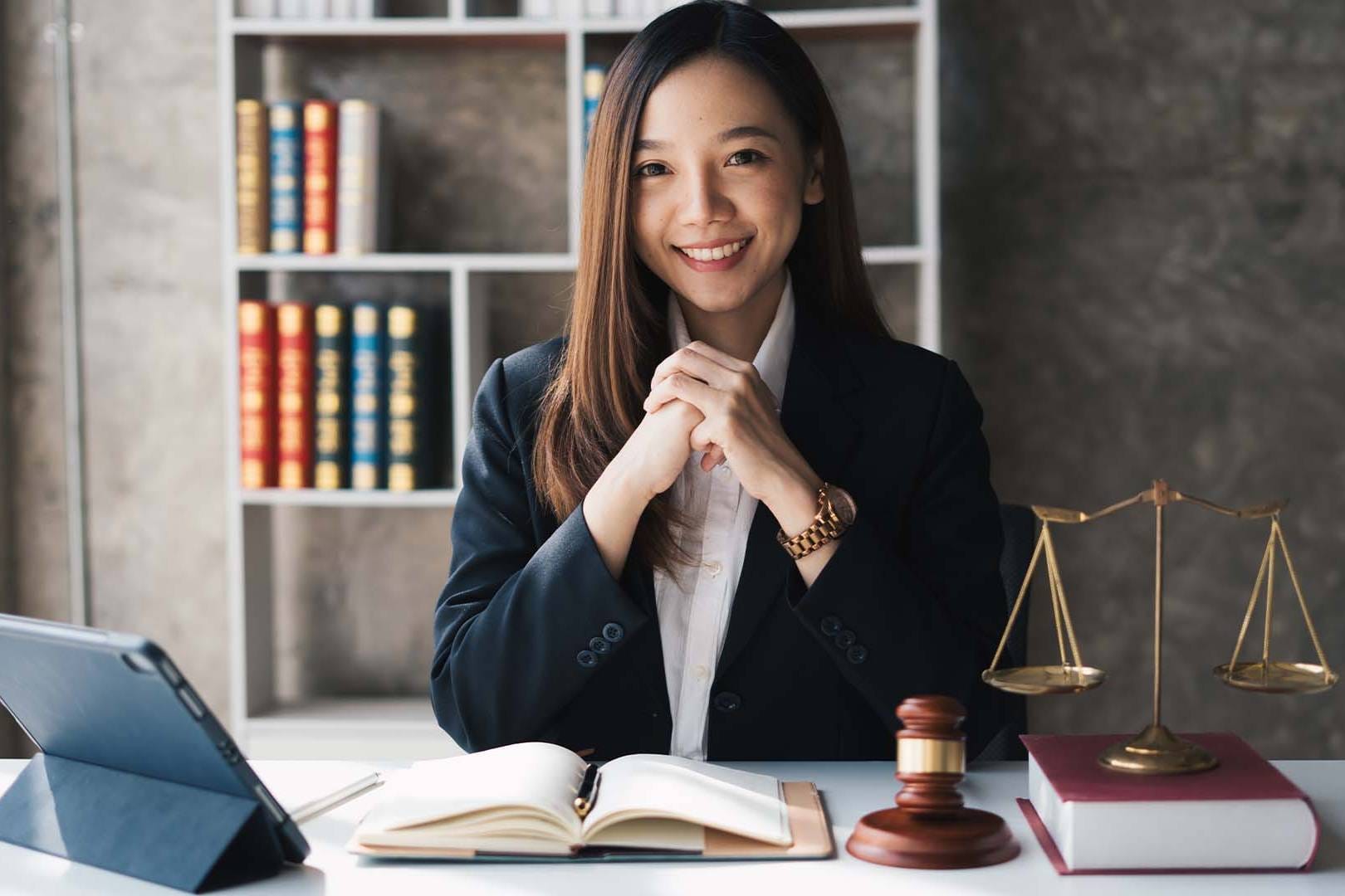 Smiling female lawyer in business suit at desk with law books, gavel, scales of justice, and tablet in professional office setting