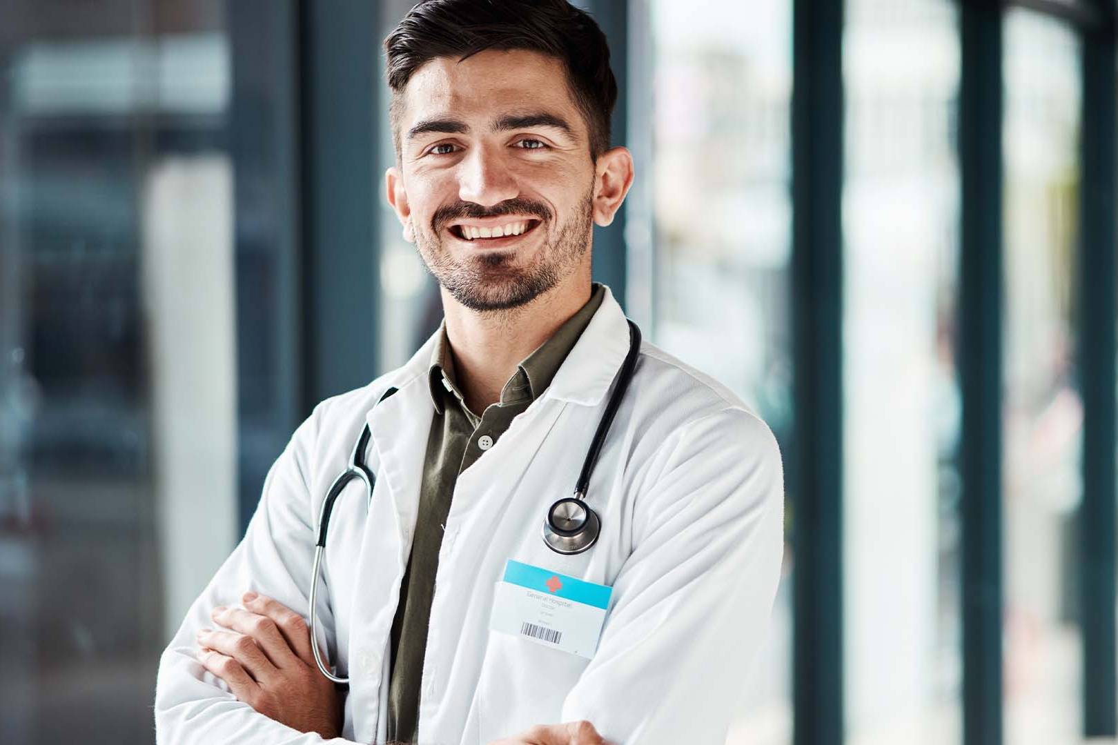 Smiling male physician in white coat with stethoscope standing in modern medical office with natural lighting