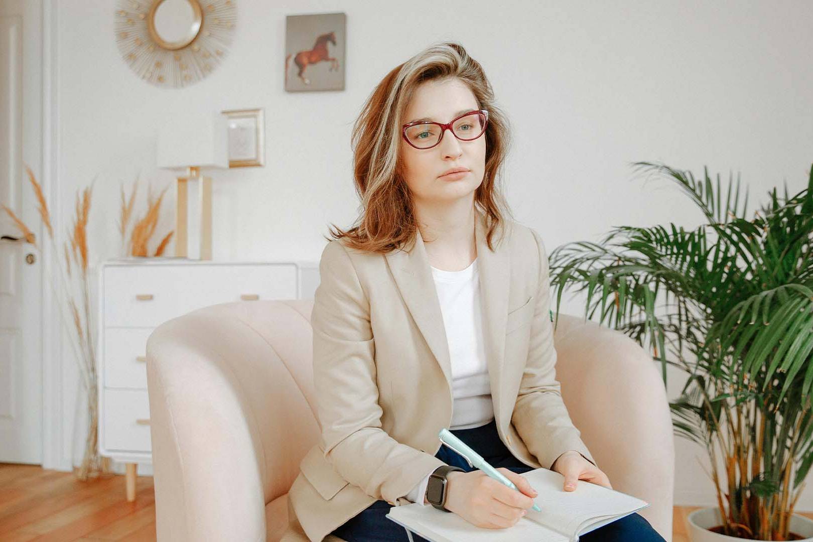 Female therapist with red glasses sitting in a beige armchair taking notes in a notebook