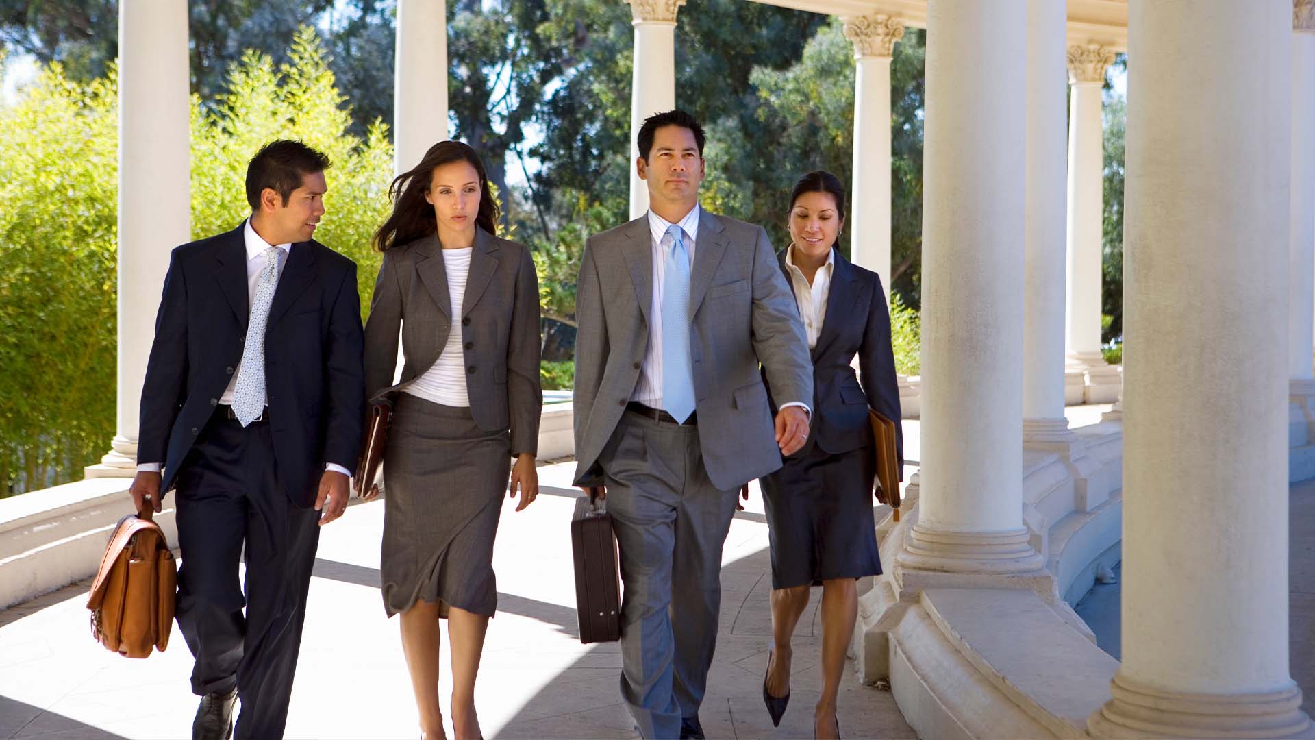 Four diverse legal professionals in business attire walking confidently through columned courthouse or law firm building entrance