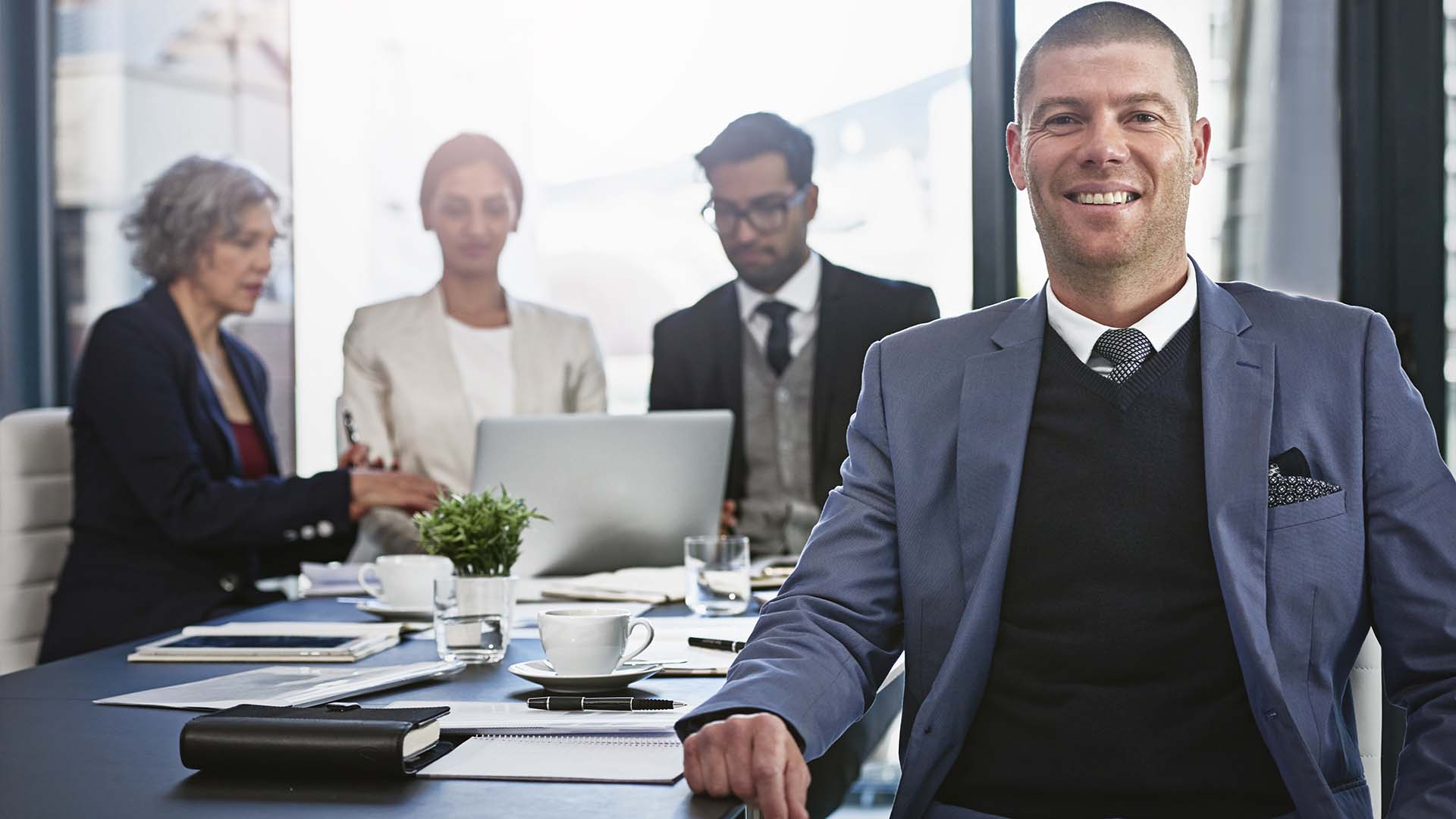 Confident businessman in a blue suit and black vest sitting at a boardroom table with colleagues working on a laptop behind him