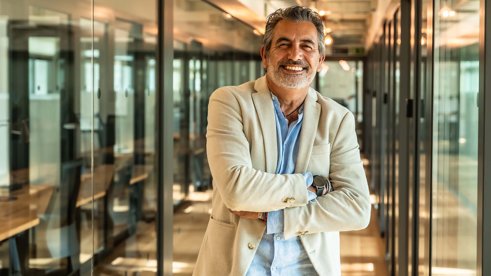 Confident mature businessman smiling with arms crossed in a modern glass-walled office hallway