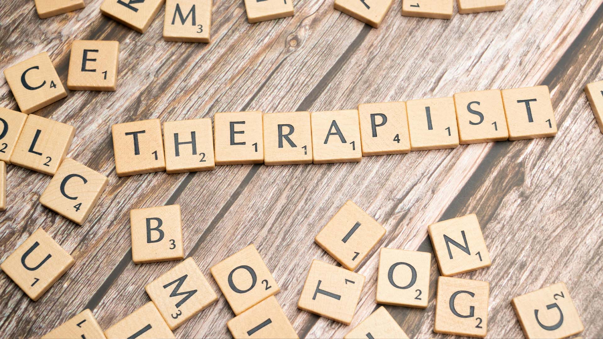 Wooden Scrabble letter tiles spelling out the word THERAPIST on a rustic wood surface with scattered tiles around it