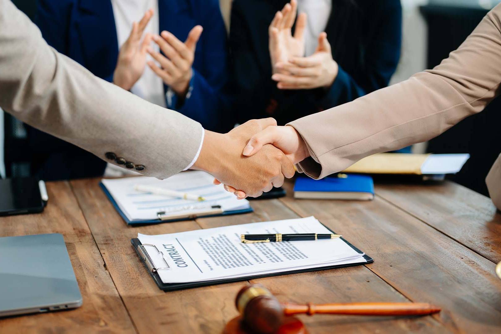 Two professionals shaking hands over a contract on a wooden desk with a gavel and colleagues applauding in the background