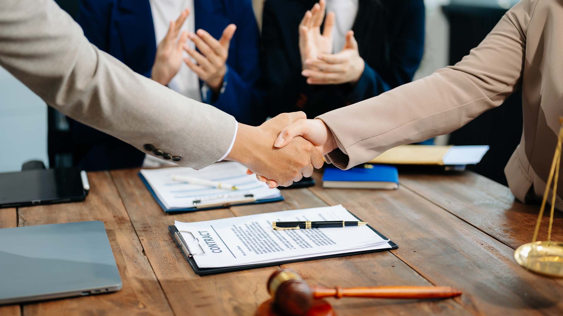 Two professionals shaking hands over a contract on a wooden desk with a gavel and colleagues applauding in the background