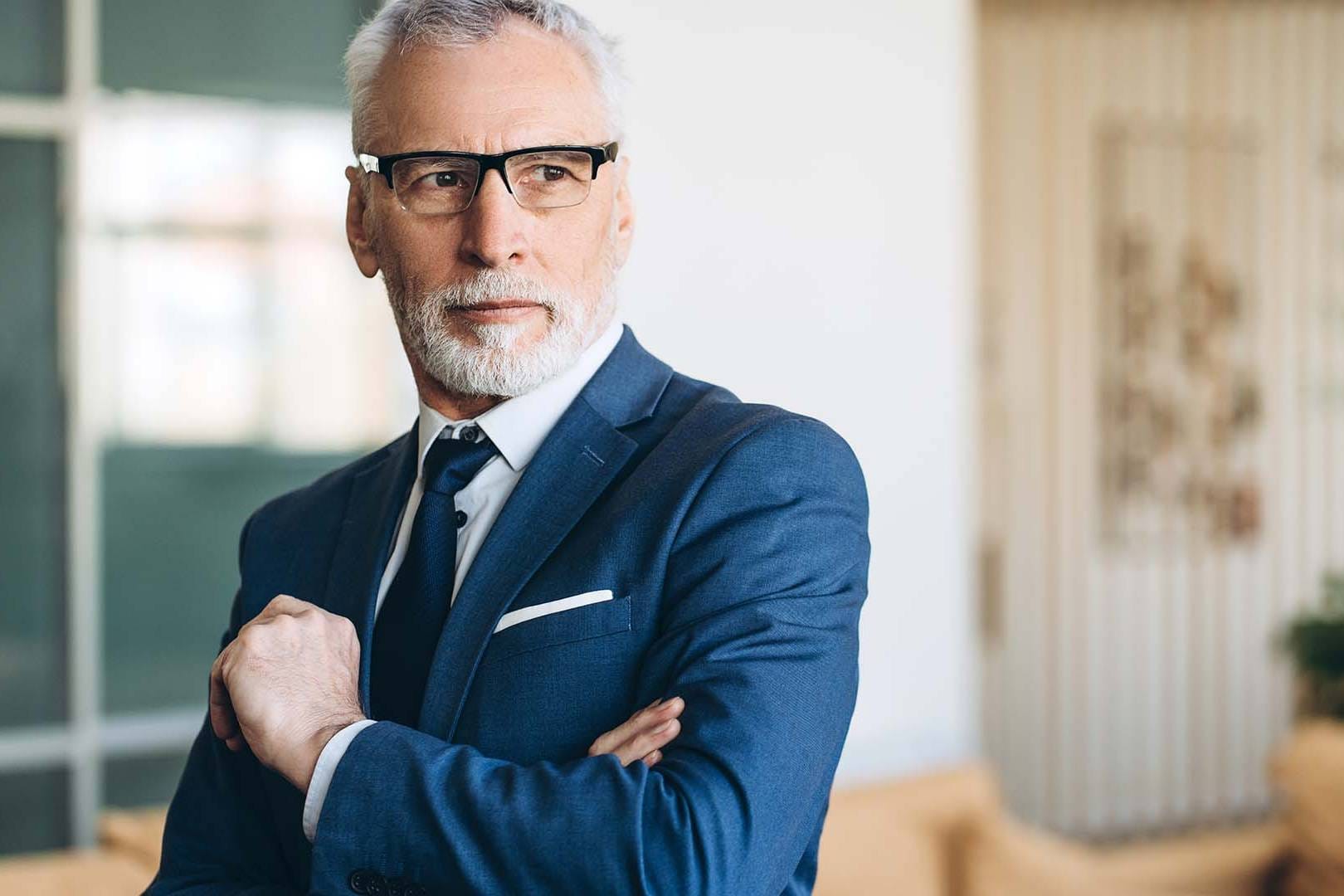 Distinguished senior executive in navy blue suit and glasses standing confidently in modern corporate office space
