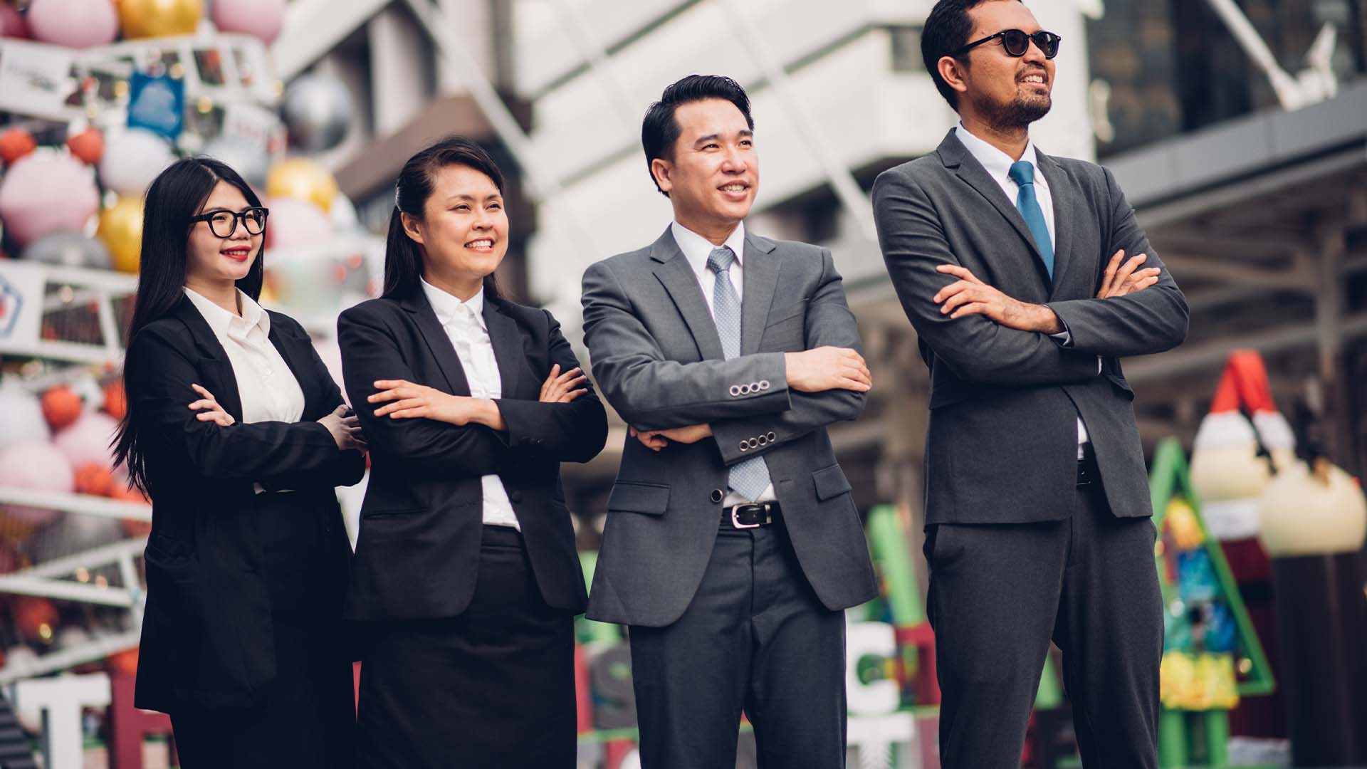 Four confident Asian business professionals in gray suits with arms crossed standing outdoors in urban setting with colorful market or festival decorations in background
