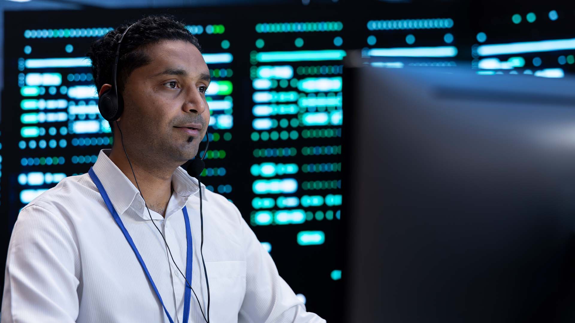 Male tech professional wearing a headset and lanyard working at a computer monitor in a server room with illuminated data screens