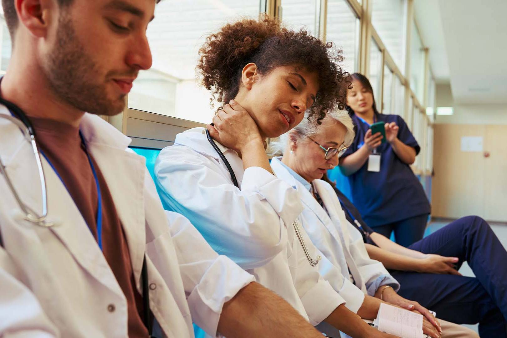 Exhausted healthcare workers in white coats and scrubs sitting in a hospital hallway showing signs of fatigue and burnout