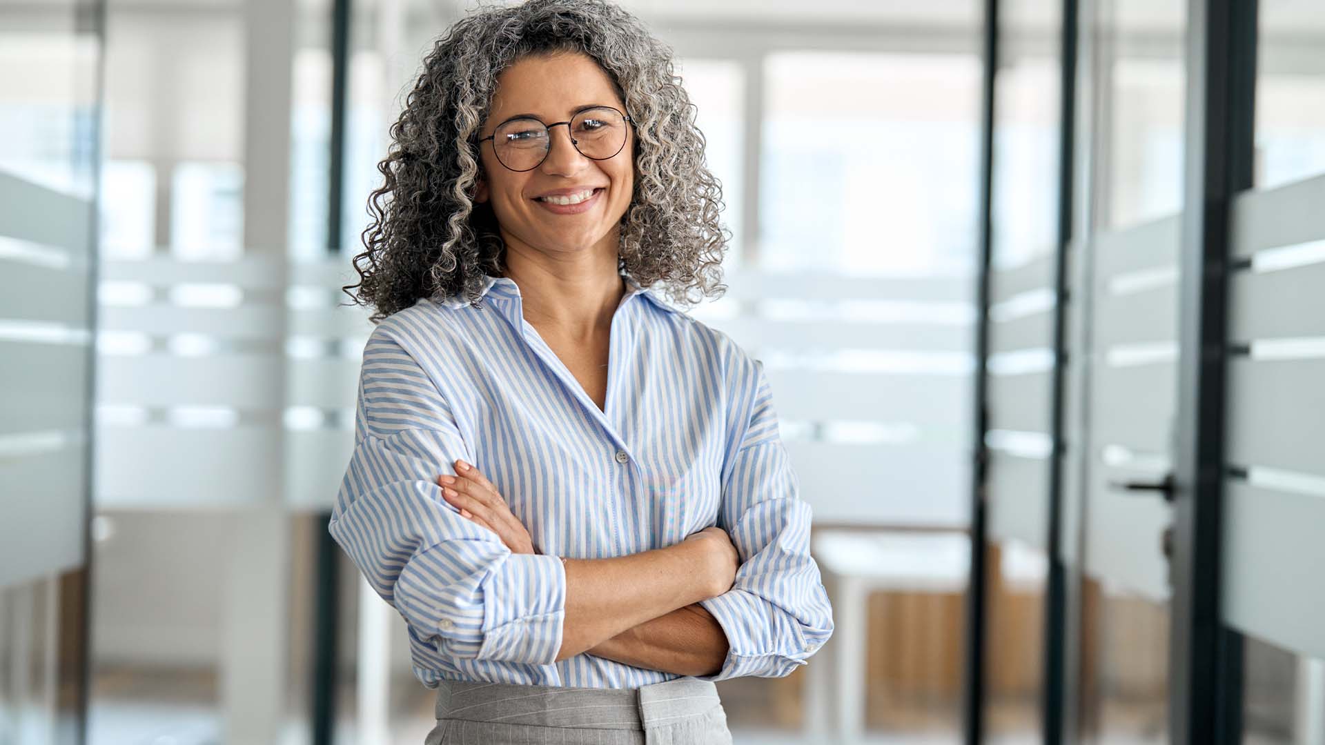 Professional woman with curly gray hair and round glasses in blue striped shirt standing confidently in modern office with arms crossed