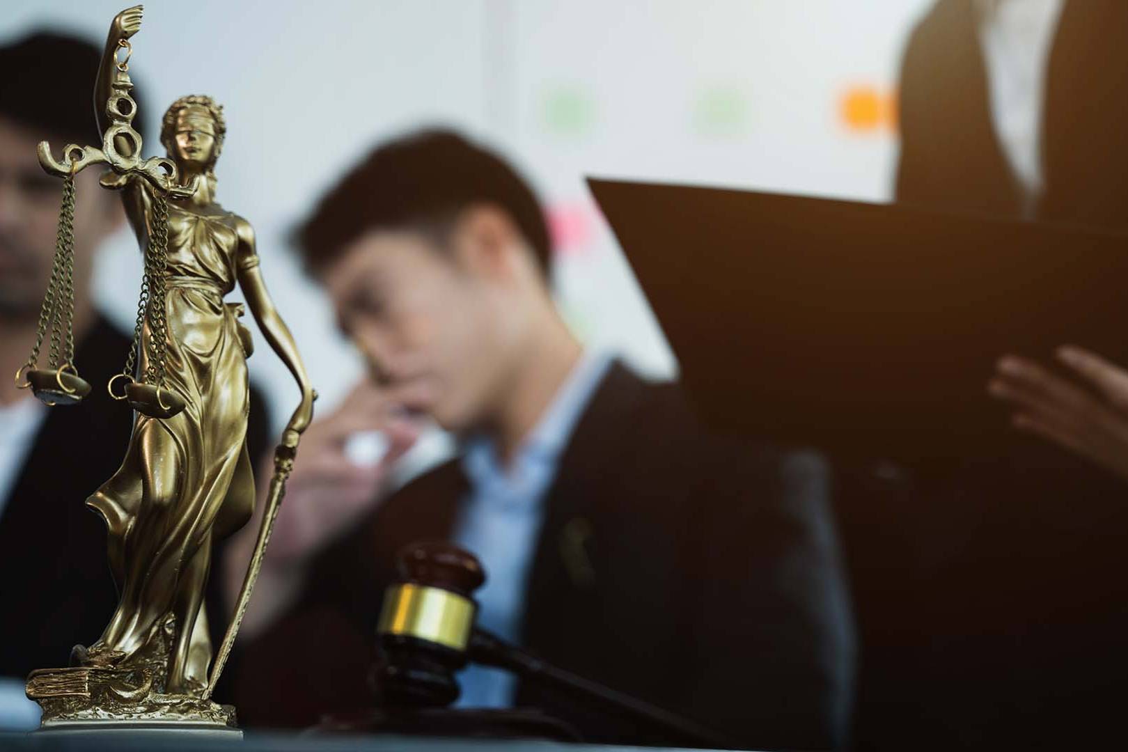 Golden Lady Justice statue with scales and sword in sharp focus on desk with blurred lawyers and gavel in background during legal meeting in modern law office