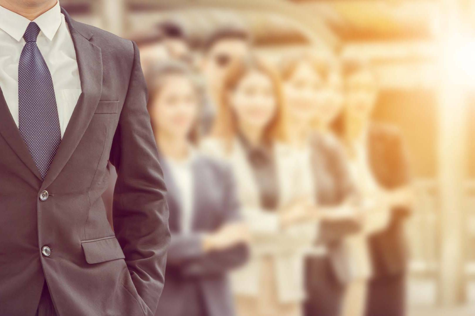 Torso of a businessman in a dark suit and patterned tie standing confidently with colleagues blurred in the background bathed in warm light