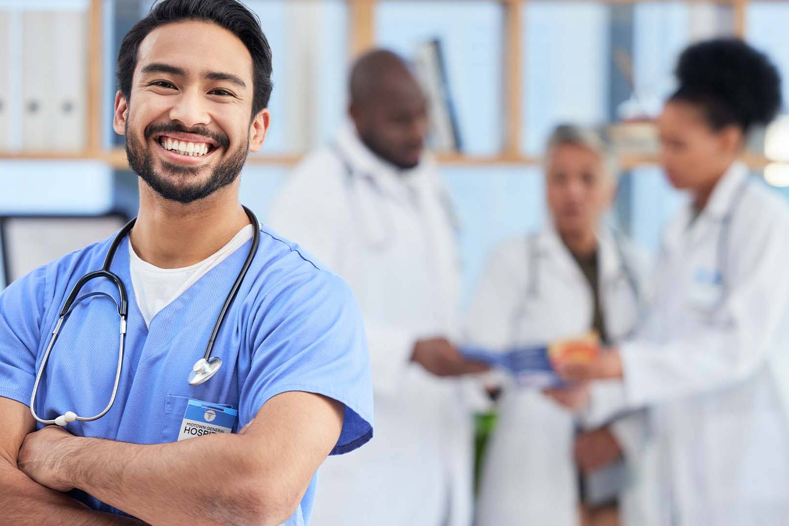Smiling male nurse practitioner in blue scrubs with stethoscope standing confidently with arms crossed in hospital setting