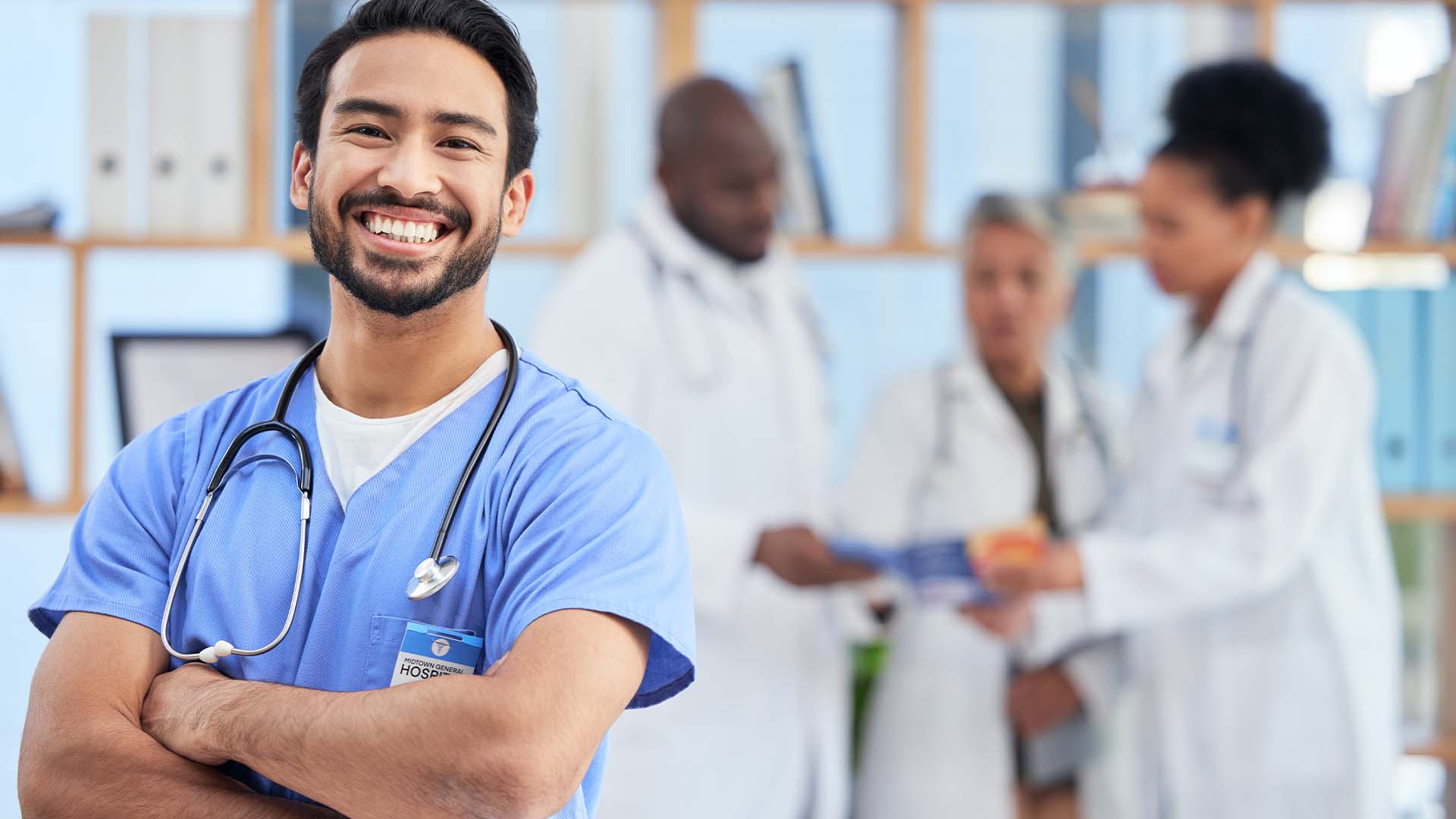 Smiling male nurse practitioner in blue scrubs with stethoscope standing confidently with arms crossed in hospital setting