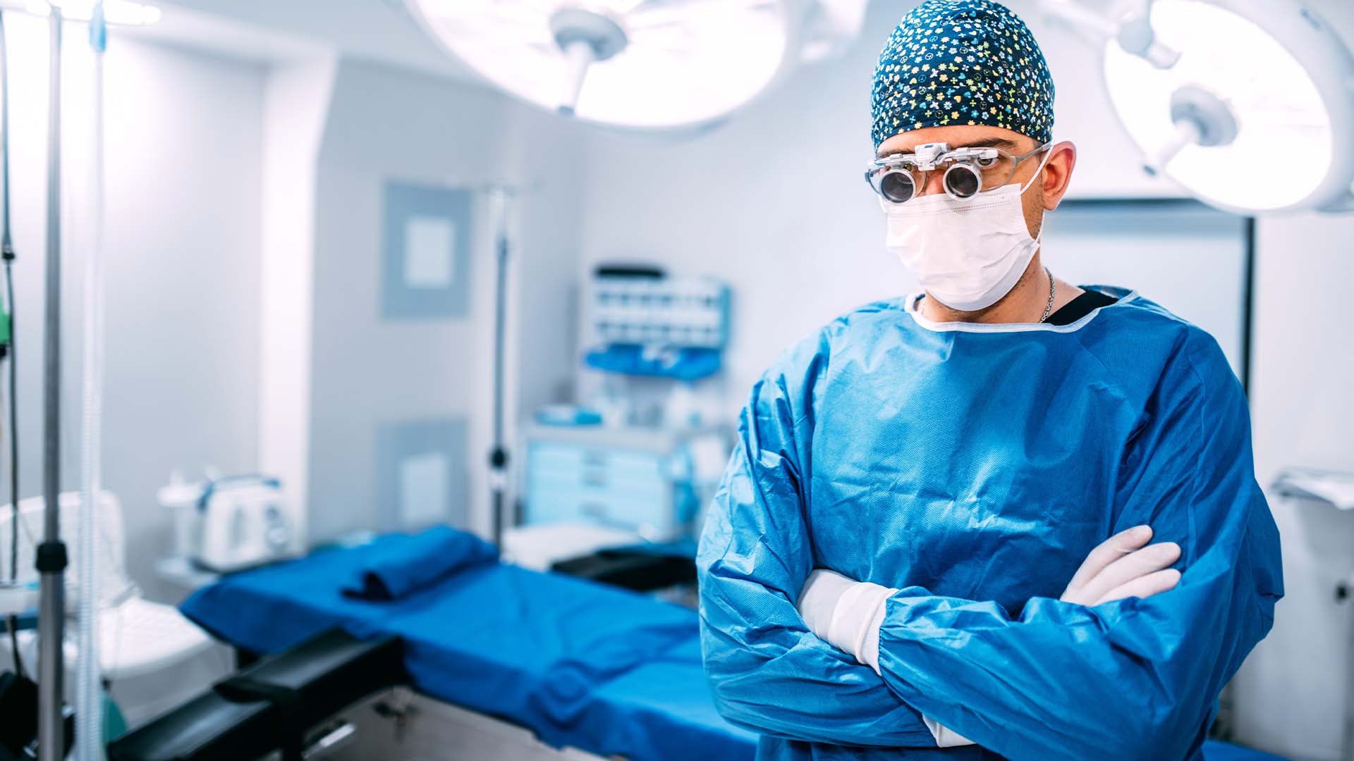 Surgeon in blue surgical gown, mask, magnification loupes, and patterned scrub cap standing in operating room with surgical lights