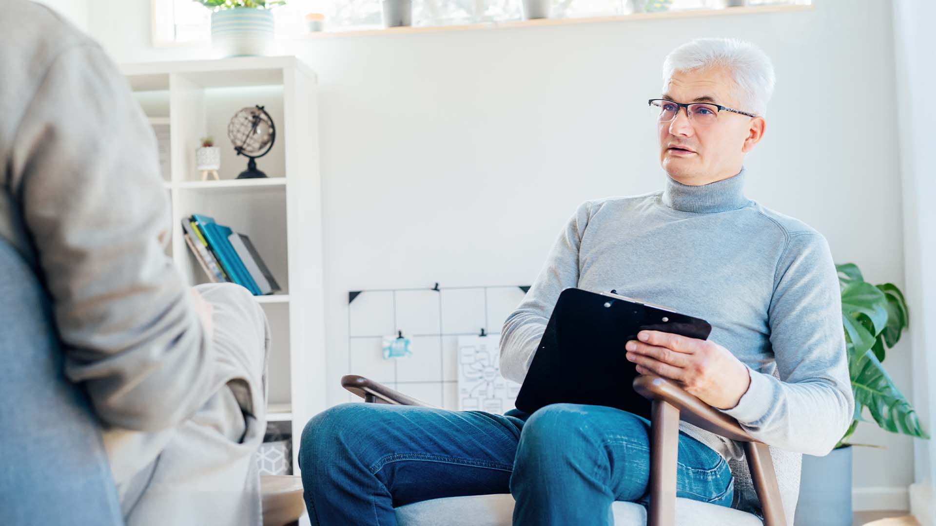 Male therapist in a gray turtleneck holding a clipboard while listening attentively during a therapy session