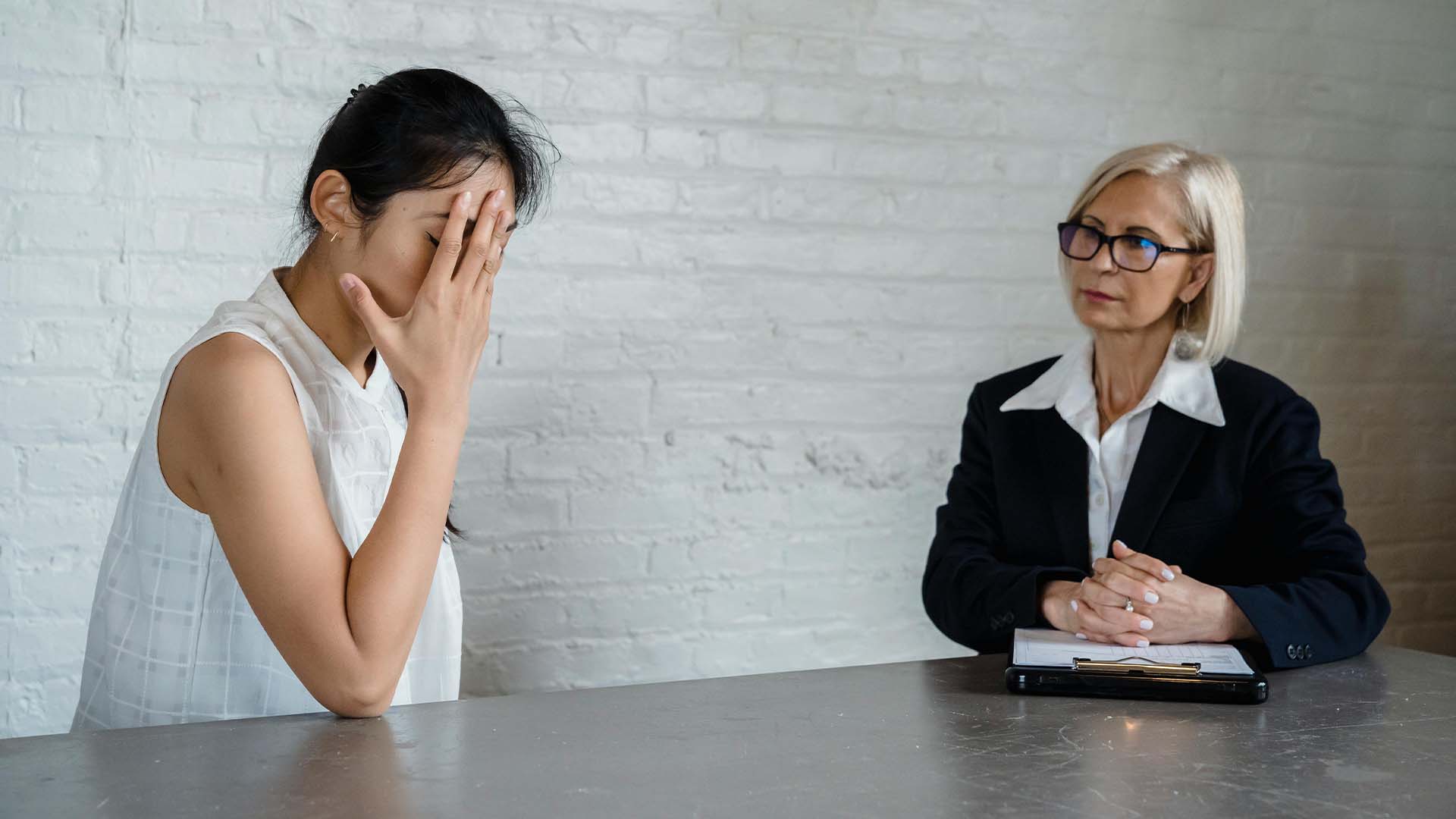 Distressed young woman covering her face while sitting across from a therapist with a clipboard at a desk