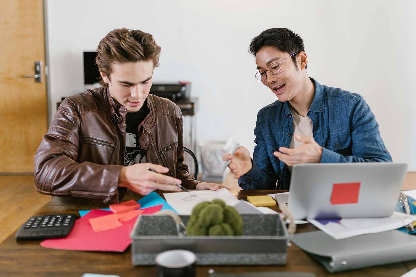 Two young men sitting at a desk collaborating over notes, sticky notes, and a laptop in a casual office setting