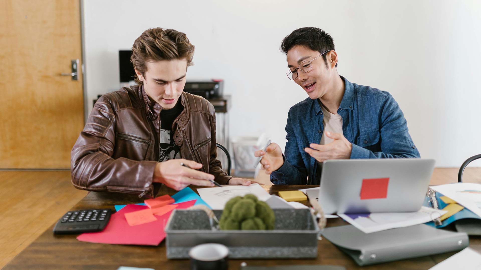Two young men sitting at a desk collaborating over notes, sticky notes, and a laptop in a casual office setting