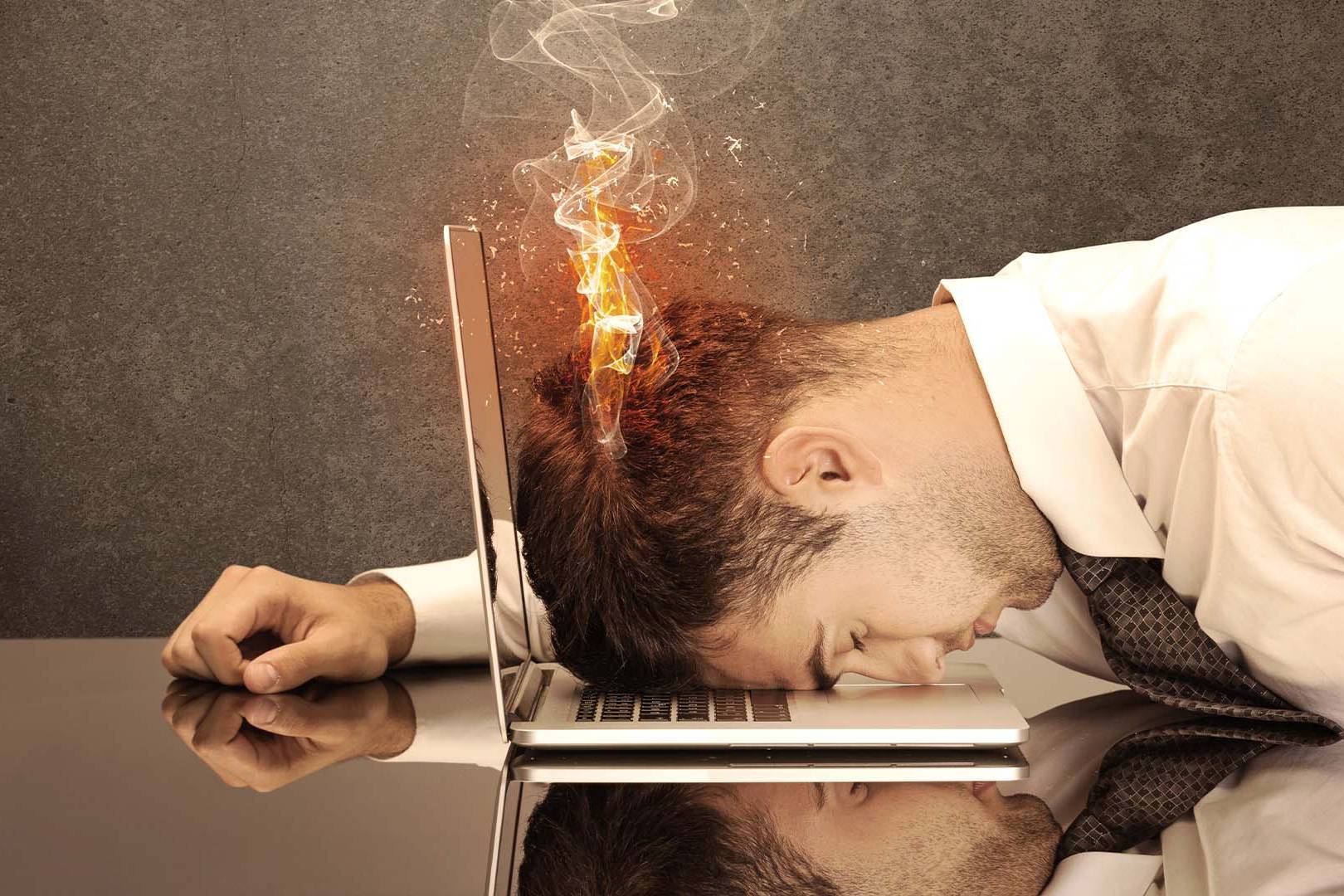 Exhausted businessman resting his face on a laptop keyboard with flames and smoke rising from his head.