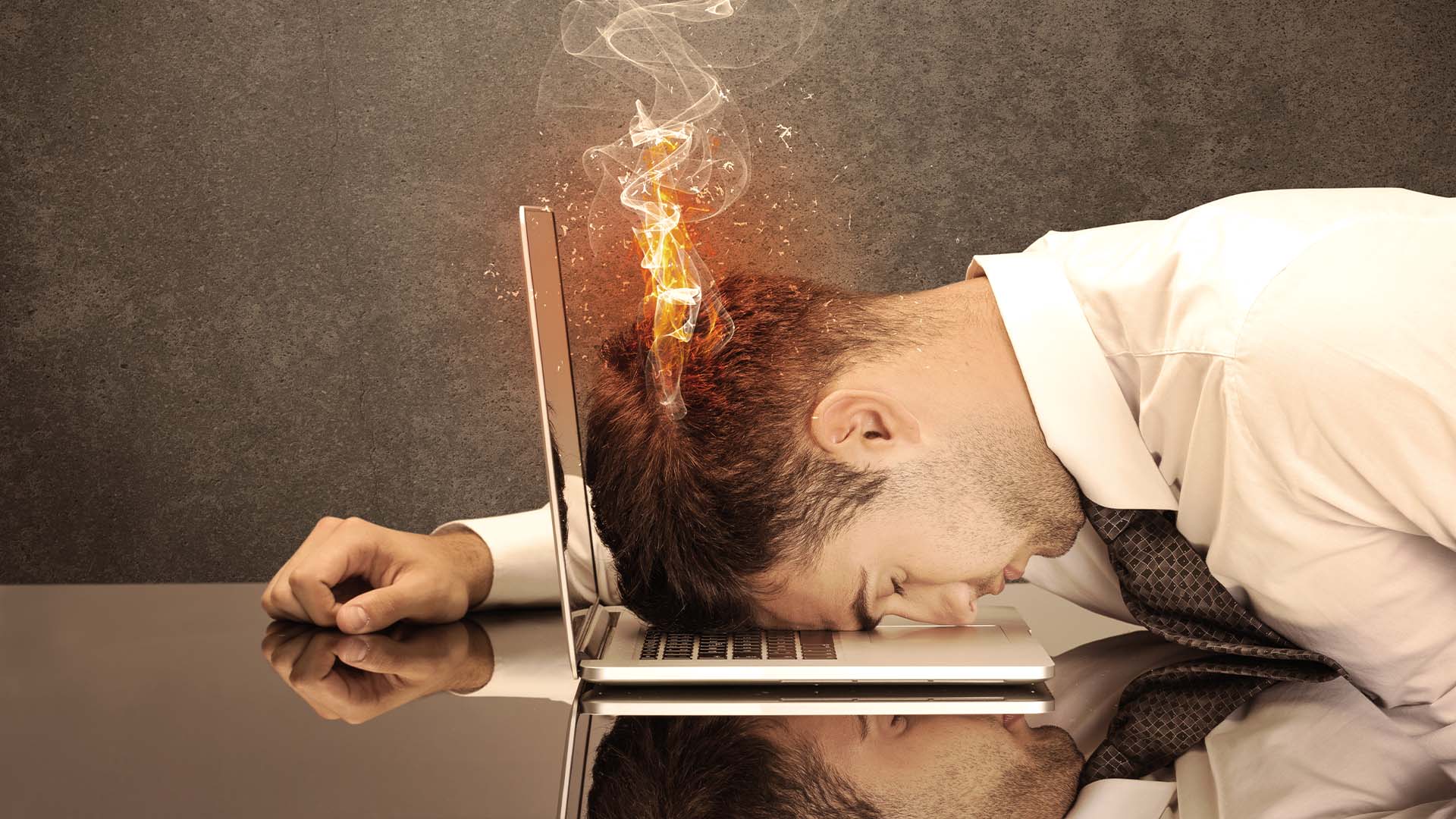 Exhausted businessman resting his face on a laptop keyboard with flames and smoke rising from his head.